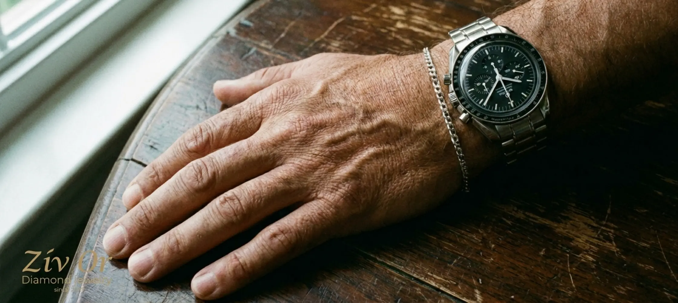 תצלום תקריב איכותי של כף יד גברית עונדת שעון כסוף אלגנטי ולצידו צמיד זהב לבן עדין מונחים על שולחן עץ כהה