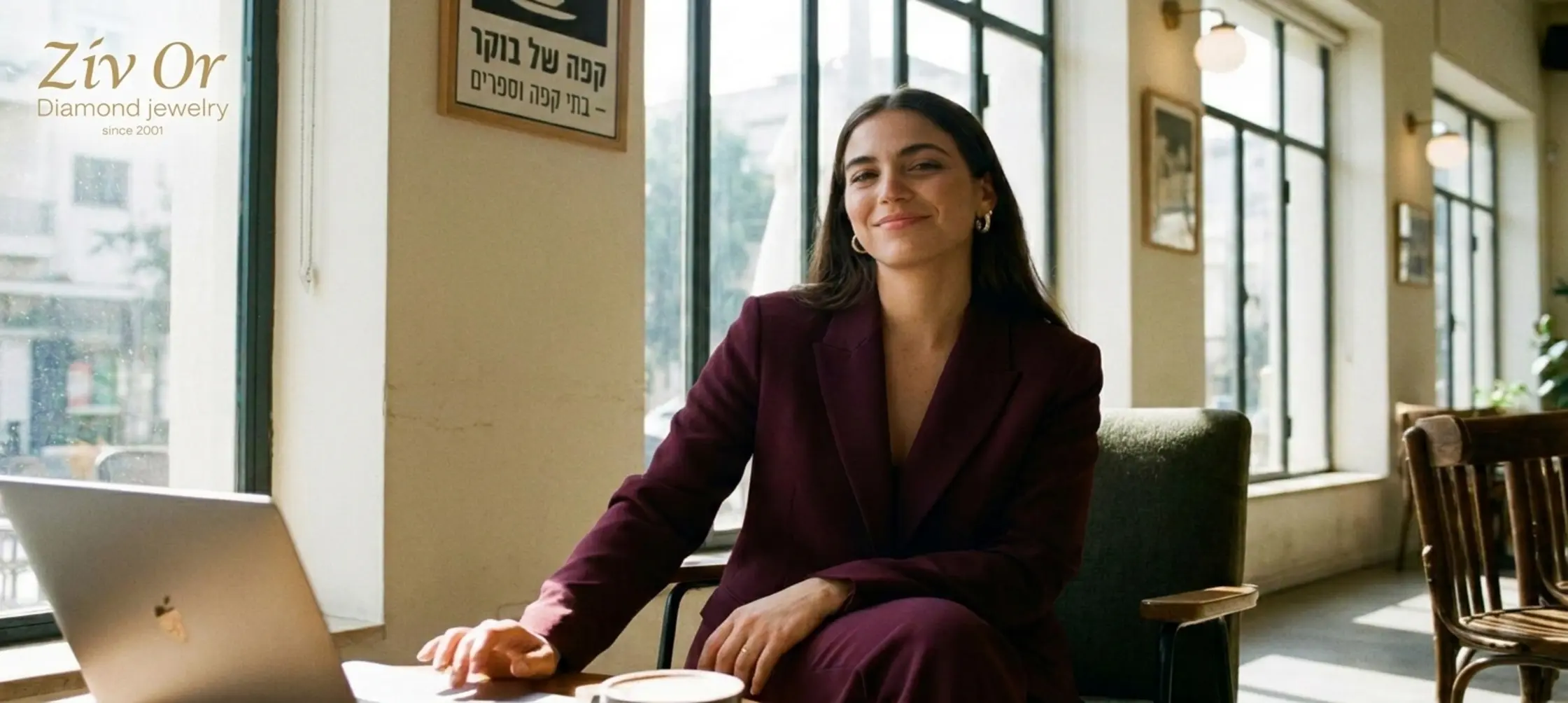 אישה צעירה ואלגנטית לבושה בחליפת עסקים מודרנית, עונדת עגילי האגיס מזהב משובצים יהלומים, יושבת בבית קפה מואר ומשדרת ביטחון מקצועי